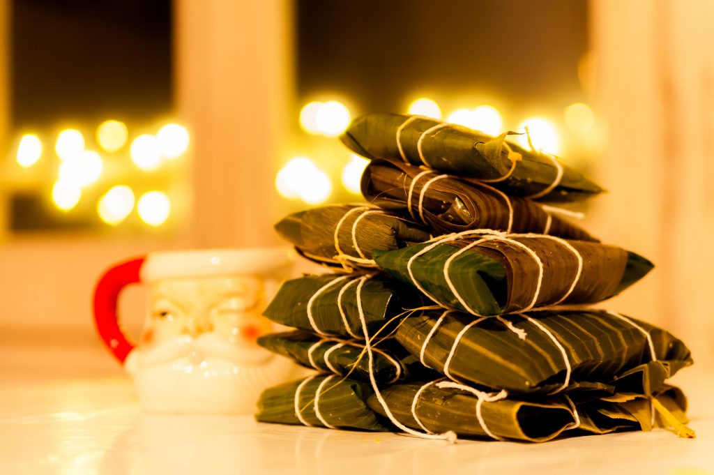 Findyello article on Caribbean Christmas recipes with image of dessert wrapped in banana leaves on a table and Christmas light in the background